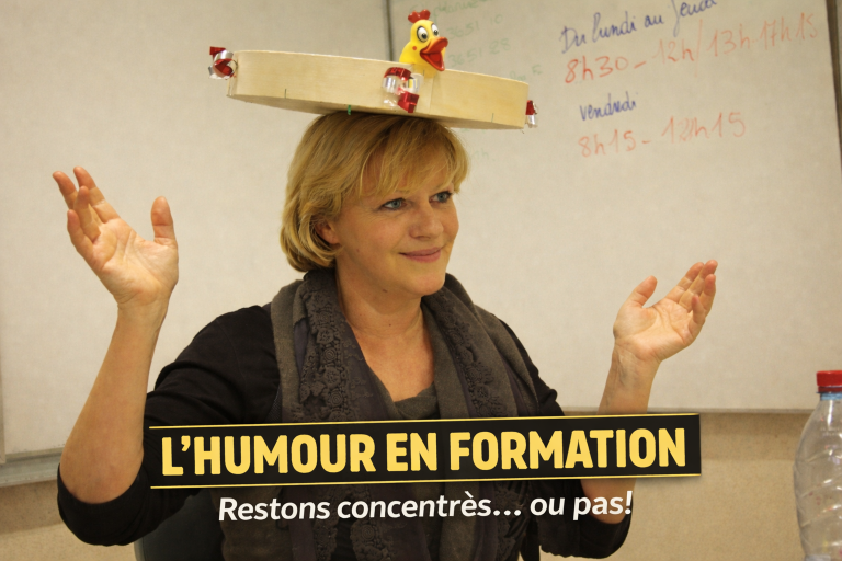 « Formatrice en salle de formation, devant un tableau blanc, portant un plateau en bois posé sur la tête. Elle a les mains levées dans un geste ouvert et un léger sourire. La scène crée un décalage visuel discret entre le cadre sérieux de la formation et un élément ludique. »